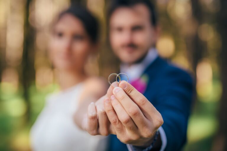 Cómo trabajo: fotógrafo bodas en Tenerife 23 Una pareja de novios muestra sus dos alianzas a cámara durante su sesión de postboda en Tenerife, con sus rostros desenfocados al fondo