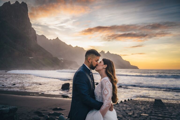 Cómo trabajo: fotógrafo bodas en Tenerife 22 Pareja de novios besándose al atardecer en una playa de arena negra de Anaga (Tenerife), con las montañas del macizo y el mar al fondo, durante su sesión de postboda