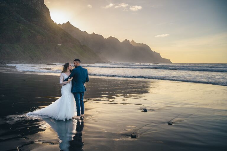 Cómo trabajo: fotógrafo bodas en Tenerife 9 Pareja de novios abrazándose en la orilla de una playa de arena negra de Anaga, en Tenerife, durante su postboda, reflejados en la arena mojada con las montañas al fondo
