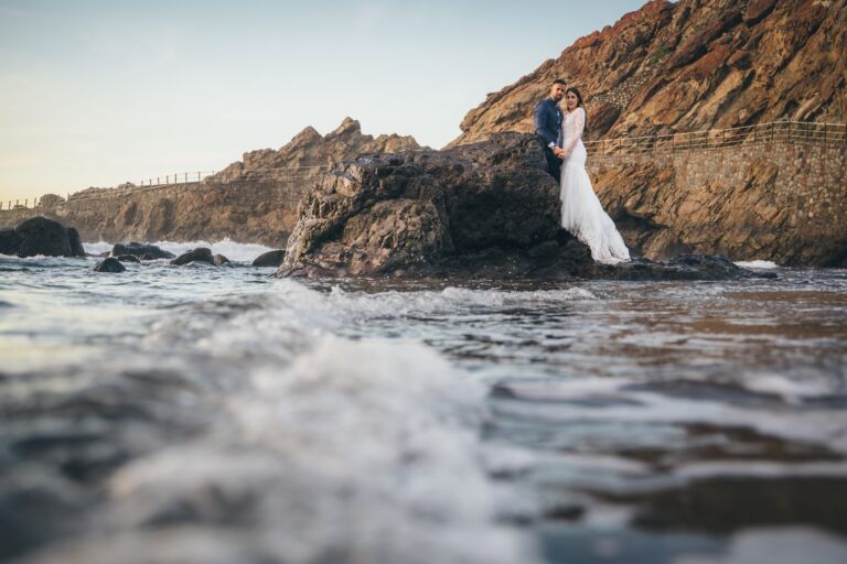 Cómo trabajo: fotógrafo bodas en Tenerife 19 Novios sobre una roca volcánica en la costa de Anaga (Tenerife) durante su postboda, con las olas rompiendo a sus pies
