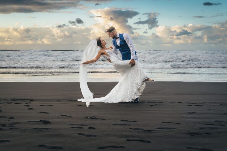Cómo trabajo: fotógrafo bodas en Tenerife 7 El novio sostiene a la novia en el aire durante su postboda en una playa de arena negra de Tenerife, con el mar embravecido al fondo
