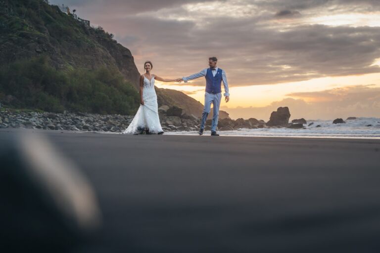 Cómo trabajo: fotógrafo bodas en Tenerife 17 Pareja de novios caminando de la mano por una playa de arena negra de Tenerife durante su sesión de postboda, con acantilados y el mar al atardecer