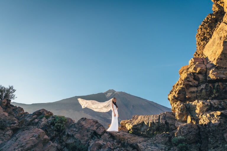 Cómo trabajo: fotógrafo bodas en Tenerife 18 Novia de pie entre rocas volcánicas del Parque Nacional del Teide durante su postboda en Tenerife, con el velo ondeando al viento y el volcán al fondo