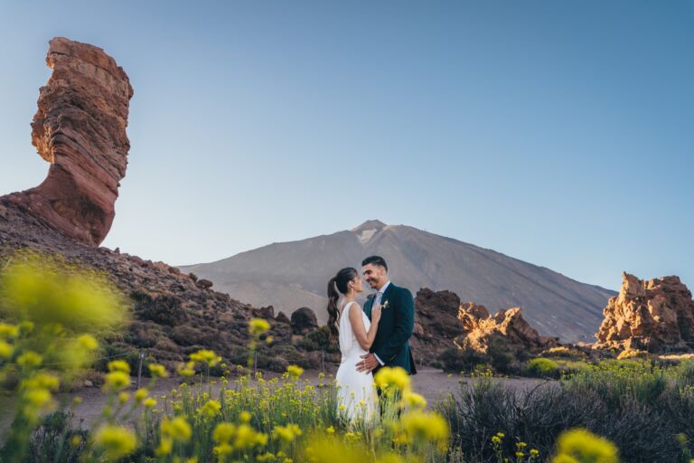 Cómo trabajo: fotógrafo bodas en Tenerife 11 Pareja de novios mirándose junto al Roque Cinchado, en los Roques de García, con el Teide al fondo y flores silvestres en primer plano, durante su postboda en Tenerife